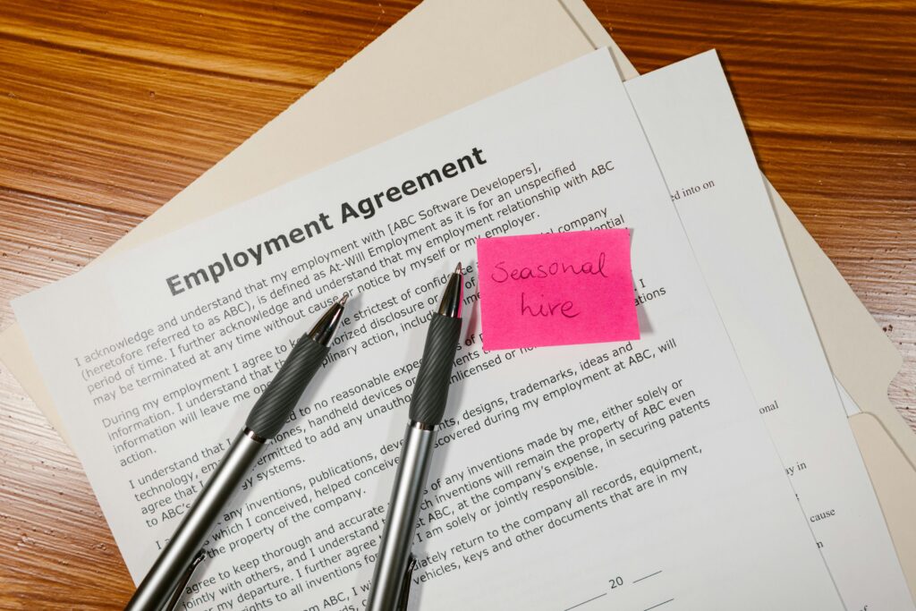 Top view of employment agreement contract with pens and seasonal hire note on wooden desk.